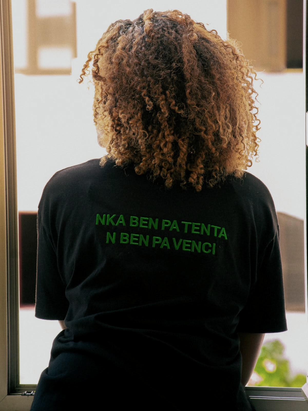 De costas, com uma t-shirt onde se lê, em kriolu, «Nka ben pa tenta, n ben pa venci».